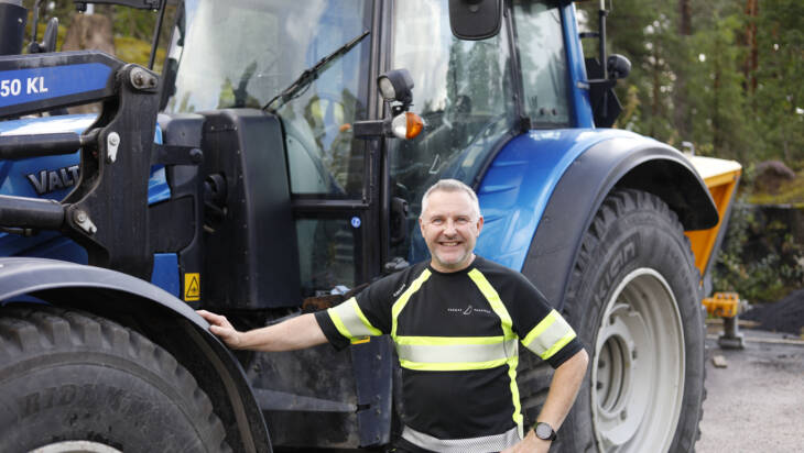 Maarakennusalan ammatti- ja erikoisammattitutkinnon suorittanut katutyönjohtaja seisoo iloisesti hymyillen kaupungin varikolla sinisen traktorin edessä.